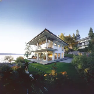 Eine moderne Villa am Ammersee mit großzügigen Glasfronten, mehreren Etagen und umlaufenden Balkonen, eingebettet in einen gepflegten Garten und gelegen direkt am Seeufer.