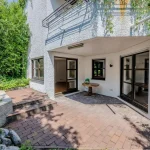 Auf dem Bild sieht man den Außenbereich eines weißen Hauses mit grober Oberflächenstruktur und großen Fenstern. Vor dem Haus befindet sich eine Terrasse mit Ziegelpflaster und einem runden Holztisch. Umliegend wachsen dichte, grüne Sträucher und Bäume.