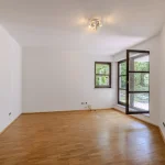 Das Bild zeigt ein leeres Zimmer mit weißen Wänden und Holzboden. Zwei Fenster und eine Balkontür führen zu einer dicht begrünten Außenfläche. An der Decke befinden sich zwei Lichtquellen. Die Adresse könnte auf einen Standort in München hinweisen.