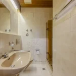 Das Bild zeigt ein Badezimmer mit beige gefliesten Wänden und einem hellen Fliesenboden. Zu sehen sind eine beige Farbene Badewanne mit einer Duschvorrichtung, eine Duschkabine mit Glastür nebenan, ein Waschbecken mit einem Spiegel darüber und diverse Badezimmerutensilien. Der Raum wird von oben durch eine beleuchtete Deckenverkleidung erhellt. Der String "Mu enchen Oldenbourgstr13a 12" scheint in diesem Zusammenhang nicht relevant.