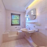 Das Bild zeigt ein Badezimmer mit pastellrosa Fliesen und Sanitäreinrichtungen. Es gibt eine Badewanne, ein Waschbecken, einen Spiegel mit Beleuchtung und ein Fenster, das auf grüne Bäume blickt. Der Raum ist hell und sauber. Der String liefert keinen relevanten Kontext.