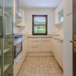 Das Bild zeigt eine leere Küche mit eingebauten beige-weißen Schränken, einem Kühlschrank, Ofen und Beleuchtung über der Arbeitsfläche. Die Küche hat ein Fenster mit Blick auf Grünflächen. Der Boden ist gefliest. Der String liefert keinen weiteren Kontext.