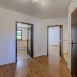 Das Bild zeigt den Innenraum einer leeren Wohnung. Wir sehen einen Flur mit Holzboden, der zu verschiedenen Räumen führt, darunter eine Küche mit weißen Schränken und Fliesenboden. Links im Bild ist ein weiterer Raum sichtbar. Die Wände sind weiß.