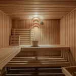 Das Bild zeigt das Innere einer Sauna aus Holz. Zu sehen sind mehrere gestaffelte Sitzbänke, ein Holzeimer mit Kelle und eine Lampe an der Wand. Es ist keine direkte Verbindung zu "Mu enchen Oldenbourgstr13a 23" ersichtlich.