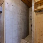 Das Bild zeigt den Innenraum einer Sauna mit angrenzender Dusche. Die Sauna aus hellem Holz hat eine geschlossene Tür und daneben ist eine offene, geflieste Dusche. Es gibt sichtbare Duschknöpfe und eine Duscharmatur an der Wand.