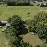 Luftaufnahme eines ländlichen Grundstücks mit grüner Wiese, Bäumen, einem kleinen Weiher, einer Trampolin, einem Gartentisch und einigen Weidezäunen. Keine direkte Beziehung zum angegebenen String "Muenchen Oldenbourgstr13a 31".