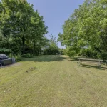 Das Bild zeigt einen grünen Garten mit einem Rasen und umgeben von hohen Bäumen. Links steht ein Grill auf einer kleinen Plattform, daneben zwei weiße Plastikstühle und ein schwarzer Müllbehälter. Rechts befindet sich ein langer Tisch mit Bänken.