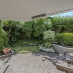 Das Bild zeigt einen gepflegten Garten einer Immobilie in der Oldenbourgstraße 13a, München. Der Blickwinkel geht von einer überdachten Terrasse aus auf dichtes Grün und einen gepflasterten Bereich. Pflanzen und Sträucher wirken gepflegt.