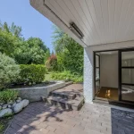 Das Bild zeigt einen überdachten Hauseingang mit einer Glastür und einem großen Fenster. Es gibt eine Terrasse mit Ziegelsteinen und Stufen, die in einen schön angelegten Garten mit Büschen, Bäumen und einem Kiesweg führen.