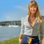 Auf dem Bild ist eine Myriam Goldhofer, Immobilienmaklerin im 5-Seenlad, Sie hat blonde Haaren und ein sympatisches Lächeln im Gesicht. Sie trägt eine graue Jacke und ein goldenes Halskettchen. Im Hintergrund sind Segelboote und der Ammersee. Das Bild lässt vermuten, dass sie gerade in dieser Region als Immobilienmaklerin tätig ist.