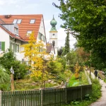 Das Bild zeigt eine malerische Straße in Obermenzing mit einem Fußweg neben einem kleinen Kanal. Links sind grüne Wohnhäuser, rechts Bäume. Im Hintergrund ragt ein Kirchturm empor, eingebettet in die herbstliche Natur. Ideal für die Suche nach einer Wohnung.