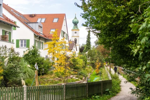 Idyllisches Wohnen in Obermenzing - jetzt kaufen! Das Bild zeigt eine malerische Straße in Obermenzing mit einem Fußweg neben einem kleinen Kanal. Links sind grüne Wohnhäuser, rechts Bäume. Im Hintergrund ragt ein Kirchturm empor, eingebettet in die herbstliche Natur. Ideal für die Suche nach einer Wohnung.