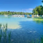 Panoramablick auf den Pilsensee mit einem Hafen, klarem Wasser und Segelbooten. Im Vordergrund Schilf, hinten bewaldete Hügel und Häuser. Ein klarer, sonniger Tag betont das idyllische, grüne Ufer.