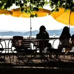 Das Bild zeigt ein Restaurant am Ufer eines Sees, vermutlich des Ammersees oder Starnberger Sees in Deutschland. Menschen sitzen unter gelben Sonnenschirmen an Tischen, genießen das Panorama und die Ruhe am Wasser.