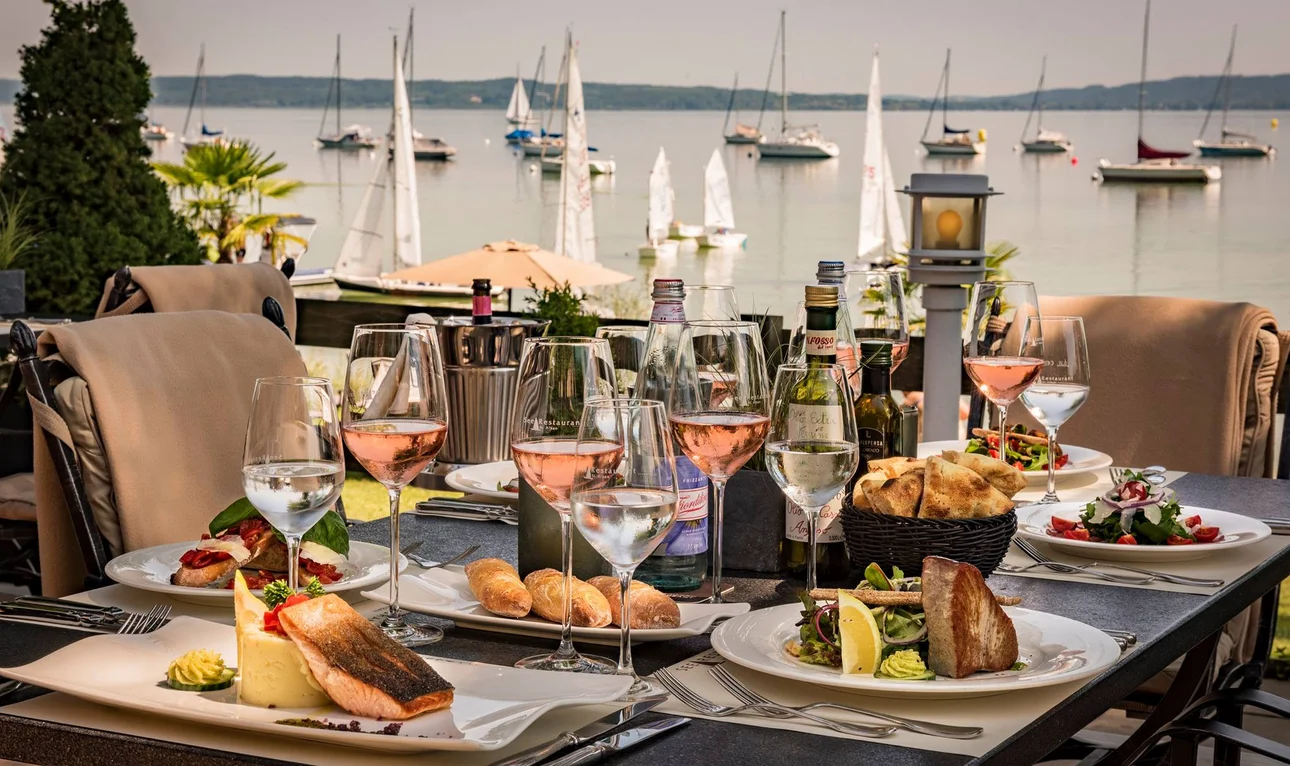 Auf dem Bild ist eine elegante Tischdekoration in einem Restaurant am Ufer zu sehen. Auf dem Tisch stehen diverse Gerichte wie gebratener Fisch, Salate und Brotkorb, ergänzt durch mehrere Gläser mit Roséwein. Im Hintergrund sind zahlreiche Segelboote auf einem See zu erkennen.