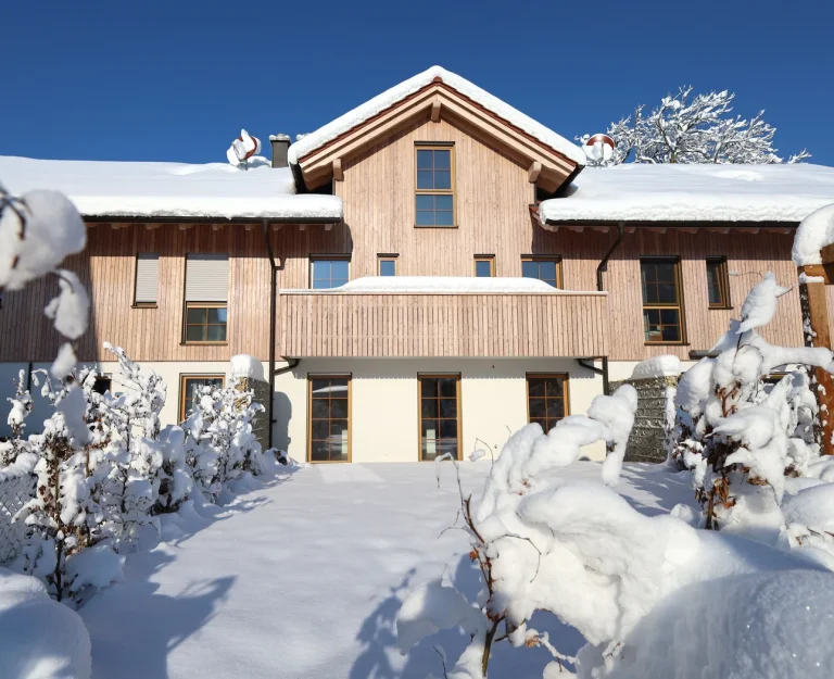 Verschneites Holzhaus unter blauem Himmel, dicht bedeckt von frischem Schnee, umringt von verschneiten Büschen.