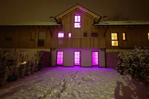 Ein großes Holzhaus unter einer Schneedecke bei Nacht. Die Fenster im Erdgeschoss leuchten pink, während die oberen ein warmes Gelb ausstrahlen. Frische Fußspuren im Schnee führen zum Gebäude.