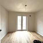 Das Bild zeigt ein leeres Zimmer mit einer weißen Wandfarbe, Holzfußboden und einer Glastür, die zum Garten führt. Der Raum wirkt unbenutzt und könnte als Schlafzimmer genutzt werden, passend zum Text "Schlafen 2 Whg. links".