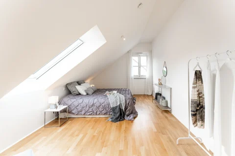 Helles Schlafzimmer unter dem Dach: Traumplatz zum Schlafen Das Bild zeigt ein Dachgeschoss-Schlafzimmer mit einem großen Doppelschlafbett, eingerichtet mit einer grauen Steppdecke und mehreren Kissen. Links neben dem Bett steht ein kleiner Nachttisch mit einer Lampe. Rechts im Raum befindet sich ein Kleiderständer voller heller Kleidung und ein Spiegel. Die Wände sind weiß, und ein großes Dachfenster sorgt für natürliches Licht.