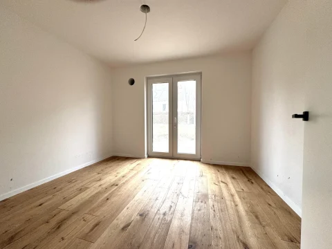 Das Bild zeigt einen leeren Raum mit beigefarbenen Wänden und einem Holzboden. Er wirkt durch eine große Terrassentür hell und könnte als Schlafzimmer oder Büro genutzt werden. An der Decke sieht man eine vorbereitete Lampeninstallation.