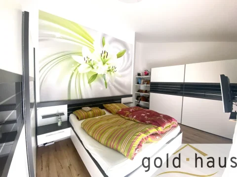 Das Bild zeigt ein modernes Schlafzimmer mit einem großen Bett, dessen Bettwäsche grün und gelb gestreift ist. Über dem Bett hängt ein großes Bild einer Lilie. Rechts steht ein großer weißer Kleiderschrank mit schwarzen Akzenten. Auf der linken Seite sind eingebaute Regale zu sehen.