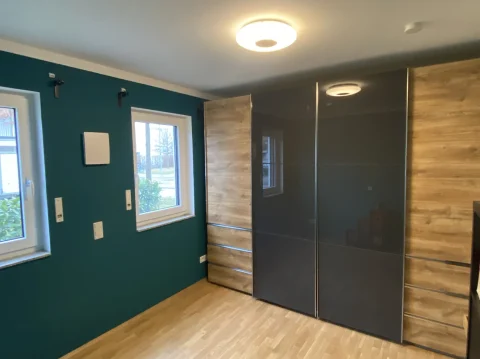 Modernes Schlafzimmerdesign in Holz und Dunkelblau Ein modernes Schlafzimmer mit türkisfarbenen Wänden, zwei Fenstern zur Straße, einem großen kleiderschrank aus dunkelgrauer Schiebetür und einer Wandverkleidung aus Holz. Auf dem Boden liegt Laminat.