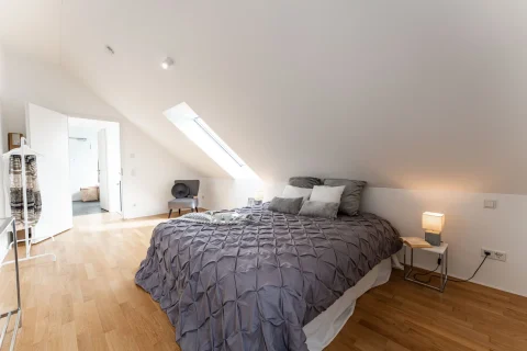 Gemütliches Dachgeschoss-Schlafzimmer mit modernem Flair Ein modernes Schlafzimmer unter einem schrägen Dach mit Parkettboden. Es hat ein großes Bett mit grauer Bettwäsche, Nachttisch mit Lampe, eine Sitzecke, eine offene Tür und Kleiderständer. Das Raumgefühl wird durch ein Dachfenster erhellt.