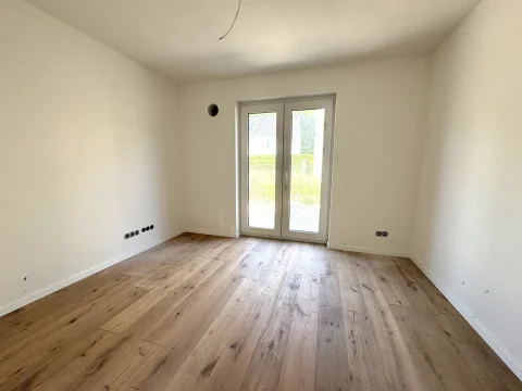 Das Bild zeigt ein leeres Schlafzimmer im Erdgeschoss eines Hauses. Es hat einen Holzboden und Wände in neutralem Weiß. Ein großes Fenster mit Terrassentür führt zum Garten. An den Wänden sind Anschlüsse für Elektroinstallationen zu sehen.