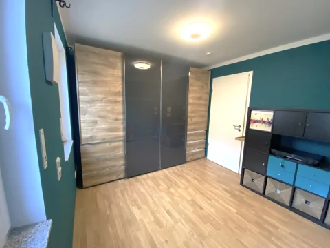 Moderne Schlafzimmereinrichtung mit großem Schrank Ein modernes Schlafzimmer mit einem großen Schrank in Holzoptik und dunklen Spiegeltüren, grünen Wänden und einem hellen Holzboden. Links sind Wandregale, rechts eine schwarze TV-Möbel. Eine Deckenlampe und eine weiße Tür sind sichtbar.