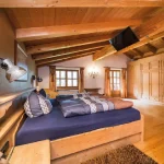 Ein gemütliches alpenländisches Schlafzimmer mit Holzbalken, Einbauschränken aus Holz, einem großen Bett mit blauen und braunen Kissen, Fenstern mit Vorhängen und einer Wandlampe über dem Bett.