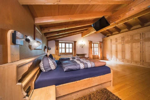 Alpen-Schlafzimmer in Tegernsee Ein gemütliches alpenländisches Schlafzimmer mit Holzbalken, Einbauschränken aus Holz, einem großen Bett mit blauen und braunen Kissen, Fenstern mit Vorhängen und einer Wandlampe über dem Bett.