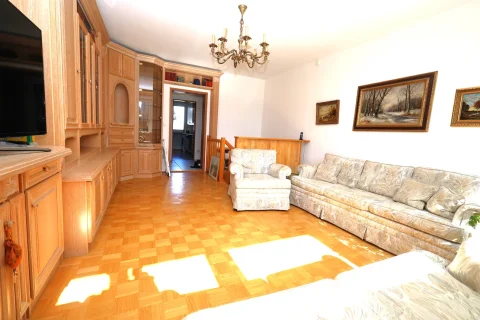 Ein helles Wohnzimmer mit Parkettboden und Sonnenlicht, das durch Fenster einfällt und Lichtflecken erzeugt. Eine Sofaecke mit zwei Couches, einem Wandregal mit TV, Bildern an der Wand und einer Deckenleuchte.