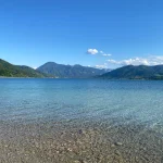 Das Bild zeigt den klaren, türkisfarbenen Tegernsee mit einem Kieselstrand im Vordergrund. Berge umsäumen den See, der Himmel ist klar mit wenigen Wolken. Ideal für entspannende Ausflüge in die Natur.