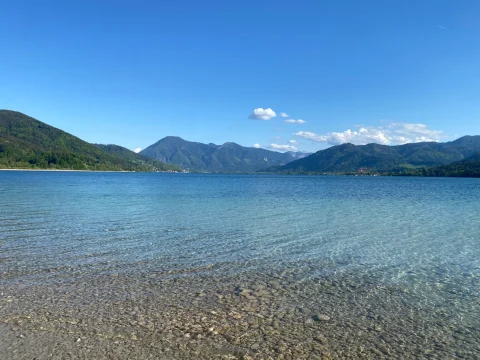 Das Bild zeigt den klaren, türkisfarbenen Tegernsee mit einem Kieselstrand im Vordergrund. Berge umsäumen den See, der Himmel ist klar mit wenigen Wolken. Ideal für entspannende Ausflüge in die Natur.