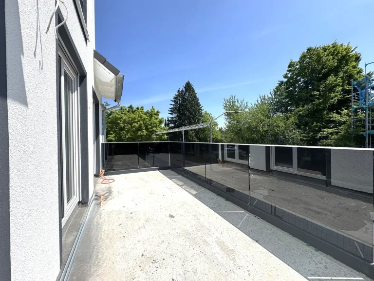 Auf der Terrasse sieht man eine graue Betonfläche umrandet von einer klaren Glaseinfriedung, welche eine offene Aussicht auf Bäume und blauen Himmel bietet. Rechts befindet sich ein Gebäude mit weißen Wänden und Fenstern.