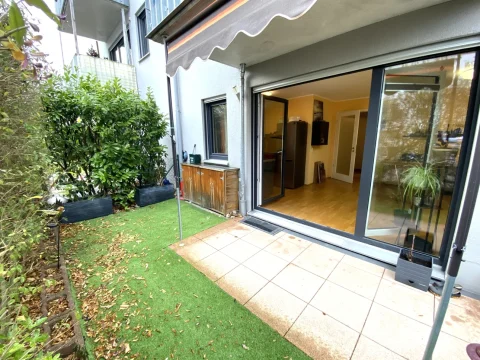 Terrasse Das Bild zeigt eine Südwest-Terrasse eines Hauses mit einer großen Glasschiebetür und einem Teil des Innenraums. Vor der Tür sind eine gepflasterte Fläche und ein Rasen zu sehen, umgeben von Pflanzen und Büschen in Pflanzkübeln.