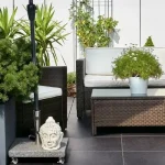 Das Bild zeigt eine moderne Terrassengestaltung mit Rattanmöbeln, darunter ein Sofa und ein Sessel. Auf einer kleinen, rollbaren Plattform befindet sich eine Buddhastatue. Pflanzen in unterschiedlich großen Töpfen und eine Kletterpflanze am Gitter ergänzen das Bild.