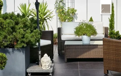 Das Bild zeigt eine moderne Terrassengestaltung mit Rattanmöbeln, darunter ein Sofa und ein Sessel. Auf einer kleinen, rollbaren Plattform befindet sich eine Buddhastatue. Pflanzen in unterschiedlich großen Töpfen und eine Kletterpflanze am Gitter ergänzen das Bild.