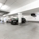 Das Bild zeigt eine helle, saubere Tiefgarage mit nummerierten Stellplätzen. Drei Autos sind geparkt. Über dem Stellplatz 28 an der Decke hängt umgekehrt ein weiteres Auto. Auf dem Boden und an einer Säule sind leichte Verschmutzungen sichtbar.