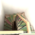 Das Bild zeigt eine Holztreppe mit Metallgeländer, die nach unten führt. Die Stufen sind mit einem bunten, gemusterten Teppich belegt, der hauptsächlich grün, gelb und rosa ist. Dies scheint die Treppe zum Hobbyraum und Bad zu sein.