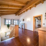 Helles Treppenhaus mit holzvertäfeltem Dach und braun gefliestem Boden. Eine hölzerne Geländer und Treppen führen hinunter. Wände mit Dekorationen, ein Kronleuchter und eine offene Tür führen in ein weiteres Zimmer.