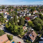 Das Bild zeigt eine Luftaufnahme eines Wohnviertels in Obersendling, München. Es ist ein Cluster von Einfamilienhäusern mit gepflegten Gärten, umgeben von Bäumen. Die Dächer sind meistens rot oder grau. Im Hintergrund ist die Skyline der Stadt sichtbar.