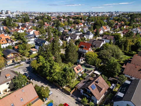 Das Bild zeigt eine Luftaufnahme eines Wohnviertels in Obersendling, München. Es ist ein Cluster von Einfamilienhäusern mit gepflegten Gärten, umgeben von Bäumen. Die Dächer sind meistens rot oder grau. Im Hintergrund ist die Skyline der Stadt sichtbar.