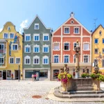 Auf dem Bild ist eine Kopfsteinpflaster-Straße in der Altstadt von Weilheim zu sehen. Rundherum stehen bunte, historische Häuser mit filigraner Architektur. Im Vordergrund steht eine steinerne Säule mit einem Blumenschmuck und Statuen.