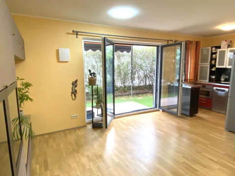 Moderner Wohn Ess Kochbereich mit Gartenblick Das Bild zeigt einen modernen Wohn-Ess-Kochbereich. Links ist eine Küche mit Edelstahlgeräten und Holzschränken sichtbar. Der Raum hat eine Glas-Schiebetür zur Terrasse und ist mit Parkettboden ausgestattet. An der Wand hängt eine Pflanze.