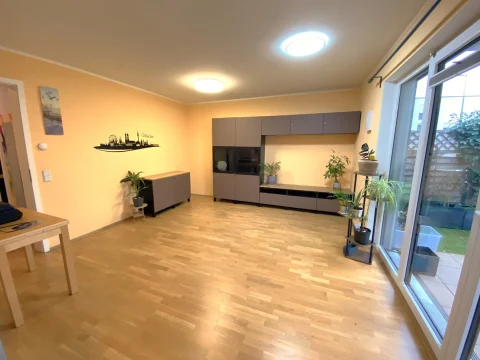 Moderner Wohn-Essbereich mit Gartenblick Das Bild zeigt einen Wohn-Essbereich mit hellen Wänden und Parkettboden. Links steht ein kleiner Tisch und ein Sideboard, rechts führt eine Glasschiebetür zum Garten. Die Küche hat dunkelgraue Fronten und eine durchgehende Arbeitsplatte. Pflanzen dekorieren den Raum.