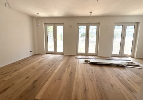 Das Bild zeigt einen leeren, hellen Wohn-Essbereich mit Holzfußboden. Drei Terrassentüren führen nach draußen. An der Decke sind Vorbereitungen für Beleuchtung erkennbar. Bauelemente liegen auf dem Boden.
