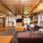 Ein gemütlicher Wohnbereich mit rustikalem Charme im Tegernseer Stil: Decke und Wandvertäfelungen aus hellem Holz, dunkelbraunes Ledersofa, Holzmöbel, Kissen, Teppiche und Fenster mit traditionellen Vorhängen.