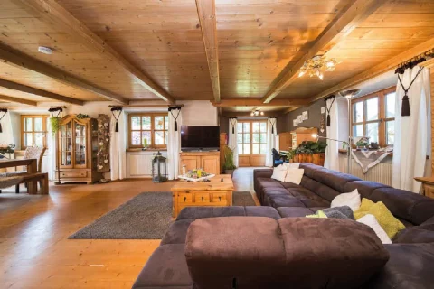 Herzstück gemütlicher Wohnbereich Ein gemütlicher Wohnbereich mit rustikalem Charme im Tegernseer Stil: Decke und Wandvertäfelungen aus hellem Holz, dunkelbraunes Ledersofa, Holzmöbel, Kissen, Teppiche und Fenster mit traditionellen Vorhängen.
