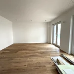 Das Bild zeigt eine leere Wohnecke mit hellem Holzboden und weißen Wänden. Zwei Türen und ein großes Fenster sorgen für viel Licht. An der Decke links sind freiliegende Kabel für Beleuchtung. Bodenleisten und Bauelemente liegen vorn rechts.